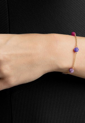 Bracelet ton or avec trois accents sphériques violet, rose et rose foncé, présenté sur une main contre un fond texturé noir.