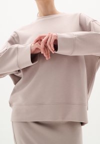 Personne portant un sweat-shirt ample beige et une jupe assortie, les mains jointes devant, sur un fond clair uni.