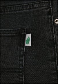 Tejido de mezclilla negra con costuras visibles. Una etiqueta verde está adjunta al bolsillo delantero, destacándose por su sutil marca.