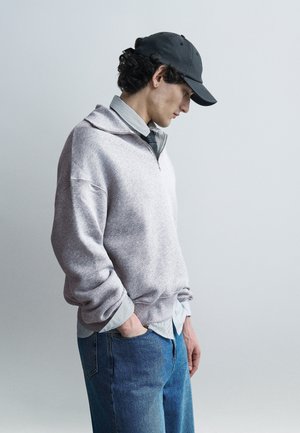 Ung mand iført en grå pullover, stribet skjorte, slips, blå jeans og sort kasket står med hænderne i lommerne og ser nedad mod en ensfarvet baggrund.