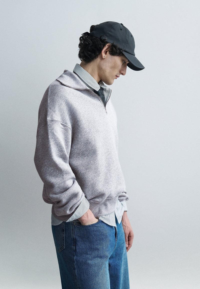 Νεαρός άνδρας με γκρι πουλόβερ, ριγέ πουκάμισο, γραβάτα, μπλε τζιν και μαύρο καπέλο στέκεται με τα χέρια στις τσέπες, κοιτάζοντας προς τα κάτω σε απλό φόντο.