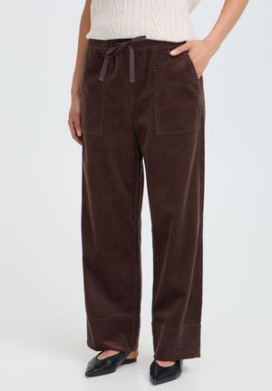 Femme portant un pantalon large en velours côtelé marron foncé avec des poches avant et une ceinture nouée, associé à des ballerines noires à bouts pointus.