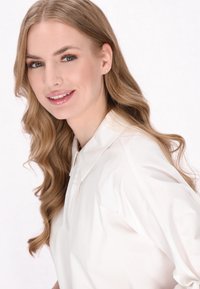 Lekka, biała koszula z kołnierzykiem, charakteryzująca się gładką fakturą i swobodnym krojem. Długie, faliste, jasnobrązowe włosy dodają delikatności wyglądowi.