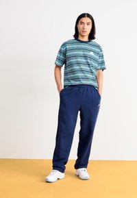 Καλοκαιρινό μπλούζα κοντό μανίκι με ρίγες σε μπλε-πράσινες αποχρώσεις με λογότυπο, συνδυασμένη με ναυτικές φόρμες και λευκά αθλητικά παπούτσια σε κίτρινο πάτωμα.