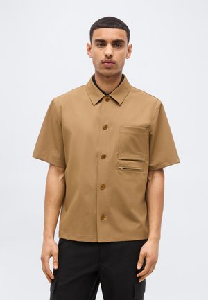 Jeune homme portant une chemise marron à manches courtes avec deux poches avant, debout devant un fond gris clair uni.