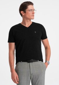 Svart t-shirt med v-ringning i mjukt tyg, med korta ärmar och en liten logga på bröstet, kombinerad med grå rutiga byxor.