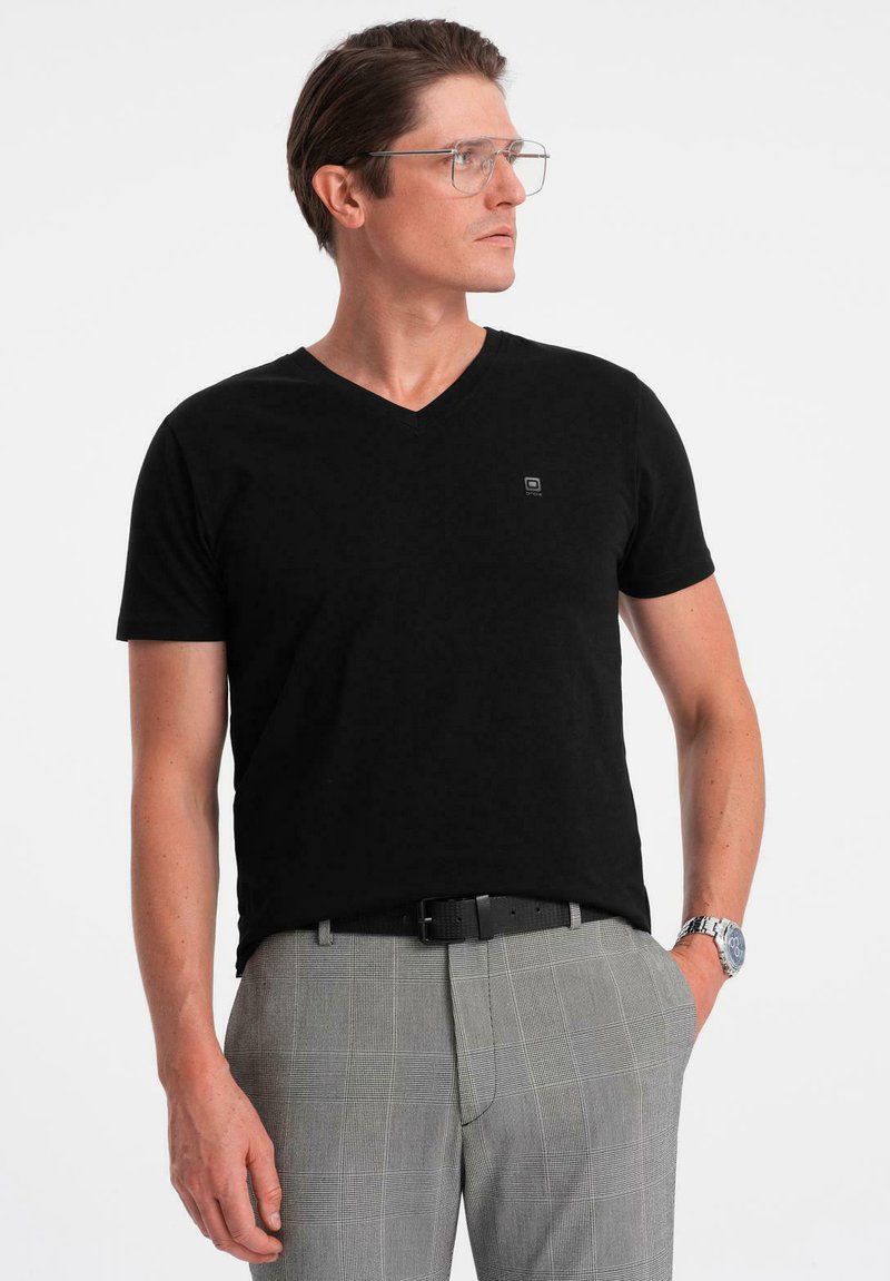 Svart t-shirt med v-ringning i mjukt tyg, med korta ärmar och en liten logga på bröstet, kombinerad med grå rutiga byxor.