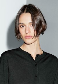 A young woman with a bob haircut wears a black button-up top, gazing directly at the camera. A soft, neutral background enhances the image.