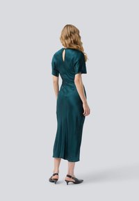 Femme aux cheveux blonds ondulés portant une robe midi en satin teal à manches courtes et des escarpins à talons kitten noirs, de dos.