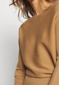 Kvinna som bär en kanelfärgad, ribbad stickad tröja med långa ärmar och en båthals, med håret delvis synligt över axeln.