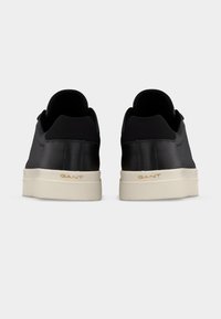 Ζευγάρι μαύρα αθλητικά παπούτσια χαμηλού ύψους με λευκές σόλες, οπτικά από πίσω, με το όνομα της μάρκας "GANT" σε χρυσό σε κάθε σόλα.
