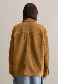 Braune Wildlederjacke mit Kragen, langen Ärmeln und Knopfakzenten. Verfügt über einen nahtlosen Rücken und eine strukturierte Oberfläche. Getragen über einem Denim-Rock.