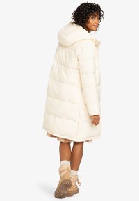 Long manteau rembourré crème clair avec capuche, présentant des coutures cousues, un ourlet droit et une silhouette carrée, accompagné de bottes beiges.