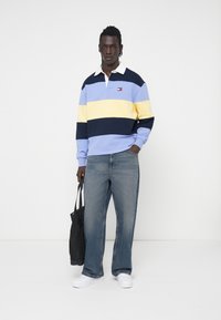 Niebiesko-żółta pasiaste koszula polo z długim rękawem i białym kołnierzykiem; noszona z jasnoniebieskimi szerokimi spodniami dżinsowymi i białymi butami, trzymająca czarną torbę.