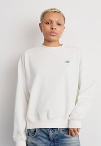 Sudadera blanca con cuello redondo, mangas largas y puños acanalados. Presenta un pequeño logo gris en el pecho. Combina con jeans de mezclilla desgastados.