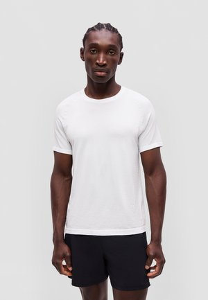 Mand med fletninger iført en enkel hvid kortærmet skjorte og sorte shorts står mod en lys grå baggrund.