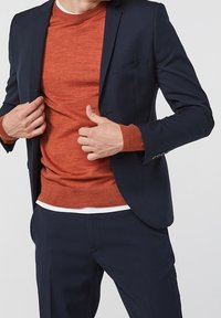 Marinblå blazer över en orange tröja med rund halsringning och ribbstickade muddar. Blazern har en bröstficka och strukturerade axlar.