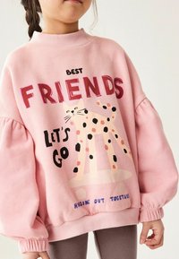 Felpa rosa con colletto alto e maniche a sbuffo, decorata con un'immagine di un gatto maculato e la scritta "MIGLIORI AMICI ANDIAMO" in caratteri grassetto.