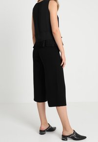 Svart ärmlös topp med plisserad fåll, kombinerad med svarta culotter. Byxorna har en lös passform; skorna har spetsiga tår med metallspikar.