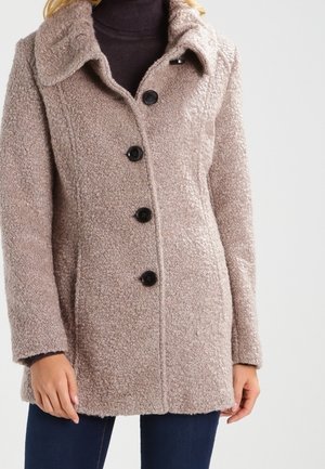 Kvinde iført en beige, tekstureret uldfrakke med sorte knapper over en mørk rullekrave, kombineret med mørkeblå jeans.