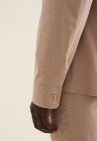 Dettaglio di una camicia a maniche lunghe di un beige chiaro, con polsini a bottoni, tessuto di cotone liscio e vestibilità rilassata. Cuciture testurizzate lungo il braccio.