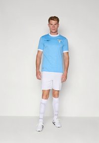 Svetlo modra nogometna majica z belimi poudarki, z logotipom kluba in kratkimi rokavi. Kombinirana z belimi hlačami in nogavicami ter črnimi kopačkami.