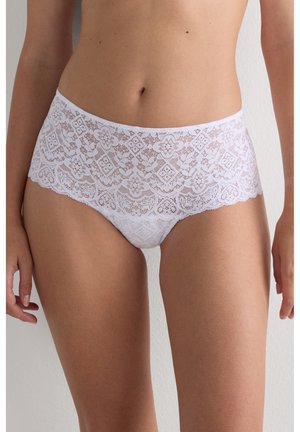 Žena nosící bílé květinové krajkové vysoké kalhotky, zobrazující střední část těla a horní stranu stehen na jednobarevném pozadí.