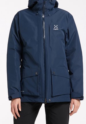 Veste imperméable bleu marine avec capuche, fermeture éclair frontale, poches sur la poitrine et à rabat, et logo Haglöfs sur la poitrine gauche.