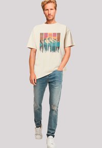 Beige grafisch T-shirt met een berglandschap en bomen, gecombineerd met lichtblauwe distressed jeans en witte sneakers. Casual pasvorm.