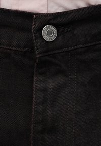 Τα μαύρα τζιν από denim διαθέτουν ίσια μέση με ασημένιο μεταλλικό κουμπί και αντίθετη ραφή. Το ύφασμα έχει λεία, ελαφρώς υφή επιφάνεια.