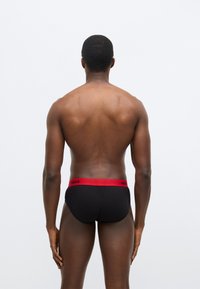 Čierne boxerky s červeným pásom, na ktorom je logo "HUGO", pohľad zozadu. Materiál pôsobí hladko a s priliehavým strihom.