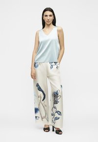 Femme portant un haut en satin bleu clair sans manches et un pantalon blanc à jambes larges avec des imprimés d'oiseaux bleus et de fleurs, des sandales noires à talons.