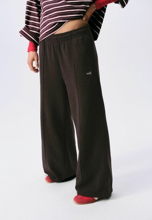 Person wearing wide-legged dark pants with "self." text on left thigh, striped long-sleeve top, and red shoes standing on white floor.