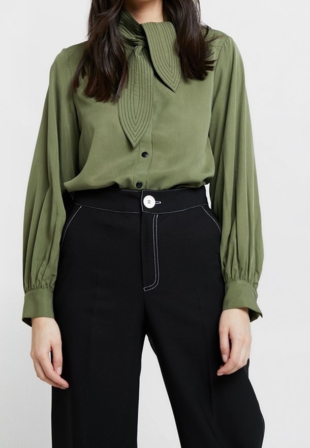 Femme portant un chemisier vert olive avec un col à nœud plissé et un pantalon noir taille haute avec un bouton blanc et des surpiqûres contrastées.