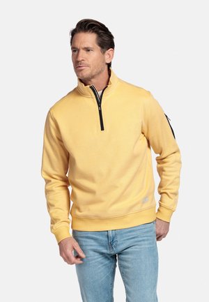 Man in een gele sweatshirt met een kwart rits en lichtblauwe spijkerbroek, staand tegen een eenvoudige lichte achtergrond.