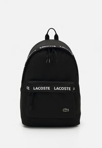 Black backpack made of fabric, featuring a rounded top, front zip pocket, and white "LACOSTE" logo strap. Lacoste logo on the front.
