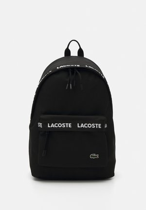 Sort rygsæk lavet af stof, med en afrundet top, frontlomme med lynlås og hvid "LACOSTE" logo strop. Lacoste logo på fronten.
