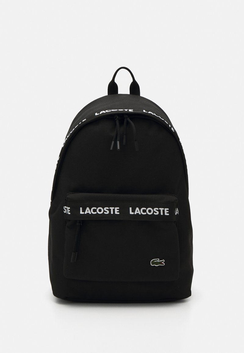 Melna mugursoma, izgatavota no auduma, ar noapaļotu augšpusi, priekšējo rāvējslēdzēja kabatu un baltu "LACOSTE" logotipa siksnu. Lacoste logo priekšpusē.
