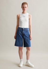 Witte geribde tanktop met blauwe tekst, gecombineerd met knielange blauwe denim shorts. Witte sneakers en enkelsokken maken de outfit compleet.