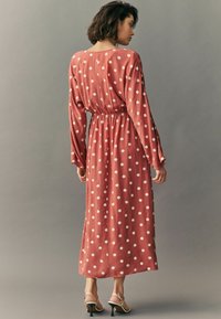 Rochie cu buline într-o nuanță de rugină, mâneci lungi, talie elastică pentru o ajustare confortabilă, material fluid cu o textură netedă și tiv până la genunchi.