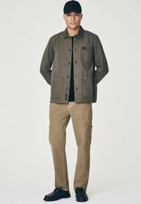 Veste en coton olive avec deux poches avant, associée à un pantalon cargo beige avec poches latérales et des chaussures noires. Casquette noire avec logo.