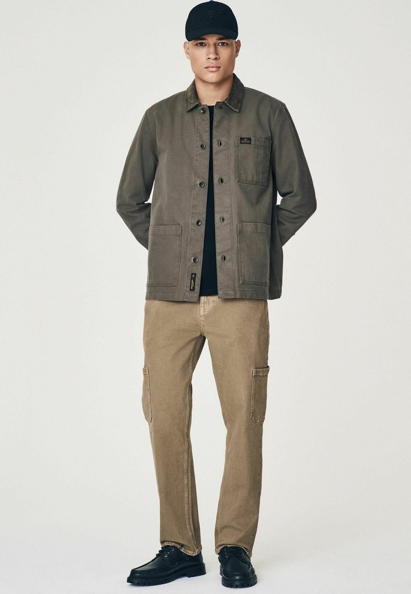 Veste en coton olive avec deux poches avant, associée à un pantalon cargo beige avec poches latérales et des chaussures noires. Casquette noire avec logo.