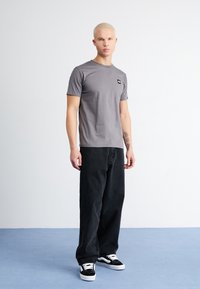Graues T-Shirt mit kurzen Ärmeln und einem kleinen bunten Logo auf der linken Brust, kombiniert mit schwarzen weit geschnittenen Jeans und schwarz-weißen Sneakern.