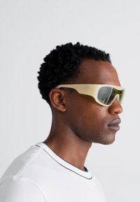 Sunglasses with a light beige frame and green-tinted lenses, featuring a sleek, angular design. Worn by a person with curly hair.