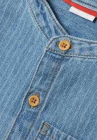 Svetlo modra denim tkanina z gumbom za zapiranje in lesenimi gumbi, vidno šivanje in žepom na prsih.