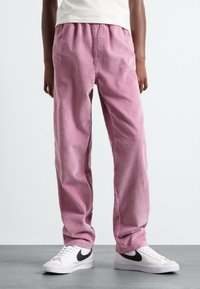 Pantalons en velours côtelé rose avec des côtes verticales, dotés d'une taille élastique. Associés à des baskets blanches, arborant un logo noir.