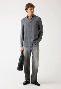 Chemise grise pour homme à boutons, manches longues, avec une poche poitrine, associée à un jean gris clair et des chaussures en cuir noires, portant un sac noir.