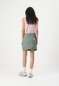 Fustă din denim verde cu două buzunare la spate și o etichetă cu logo. Asortată cu un top fără mâneci de culoare roz deschis și adidași în nuanțe neutre. Fundal alb.