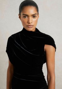 Čierne sametové šaty s priliehajúcou siluetou, vysokým výstrihom a navrhnutými ramienkami, s hladkou textúrou a elegantným leskom.