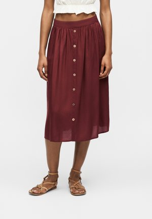 Mujer con una falda burdeos de cintura alta, con botones en la parte delantera, hasta la rodilla, sandalias marrones con tiras y una camiseta corta blanca.